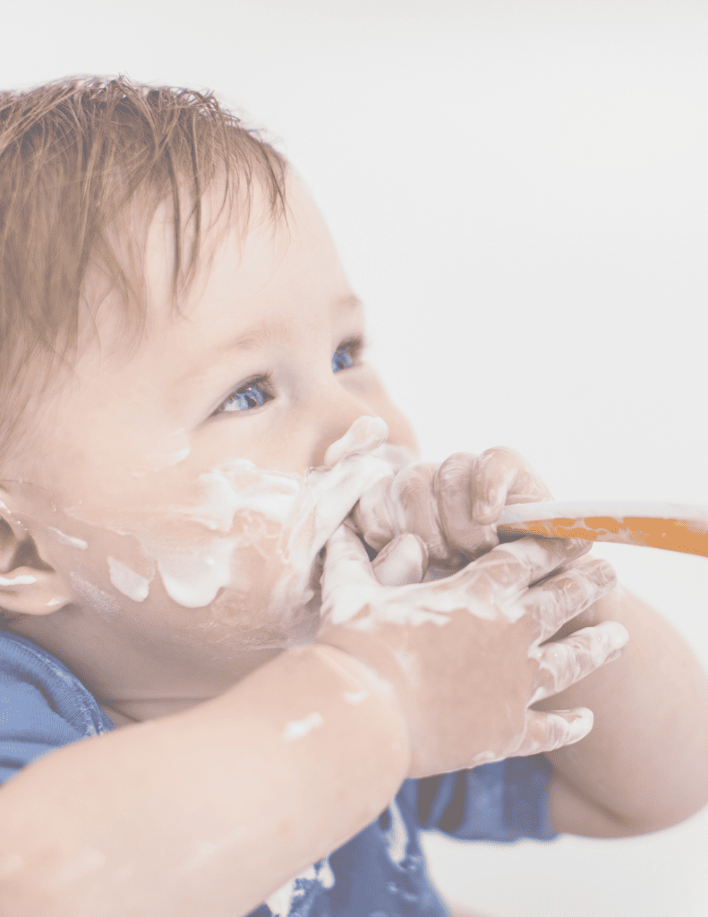 baby with orange spoon in his mouth and yogurt on his face and hands.