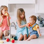 3 kids sitting with feet in the kitchen sink eating apples.