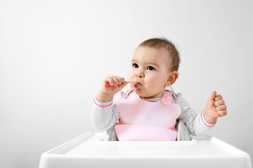 baby in pink bib in a high chair with a spoon in her mouth.
