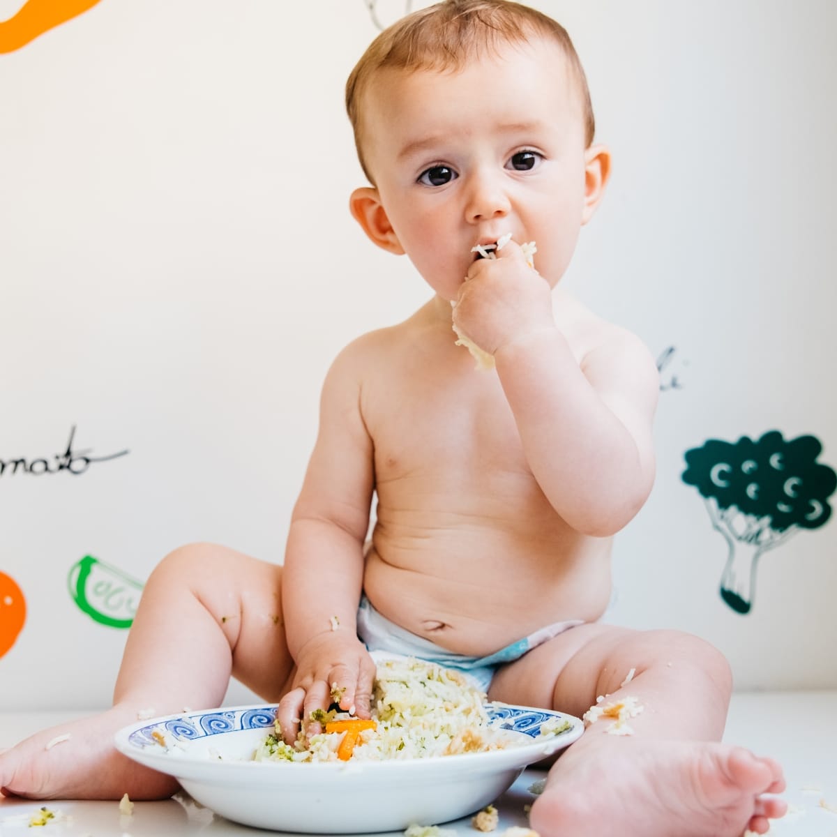 baby in a diaper sitting on the ground eating from a bowl of food with his hands.