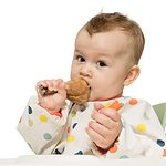 baby in a smock eating a chicken leg.