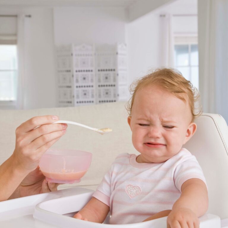 spoon of puree being offered to a baby who is refusing with a scowl on her face.