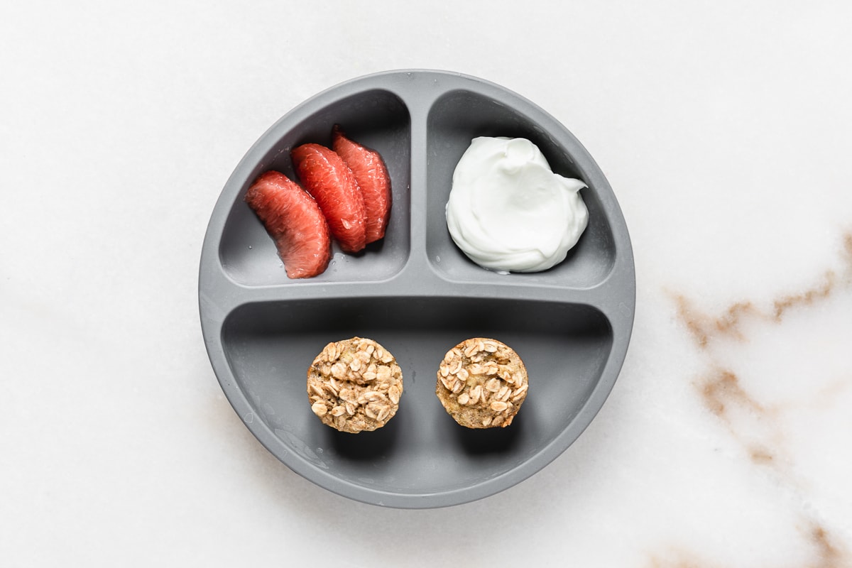 grey baby plate with grapefruit segments, yogurt, and mini banana baked oatmeal muffins on it.