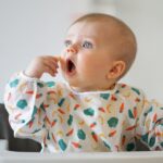 baby in a high chair and smock bringing a piece of food to his mouth.