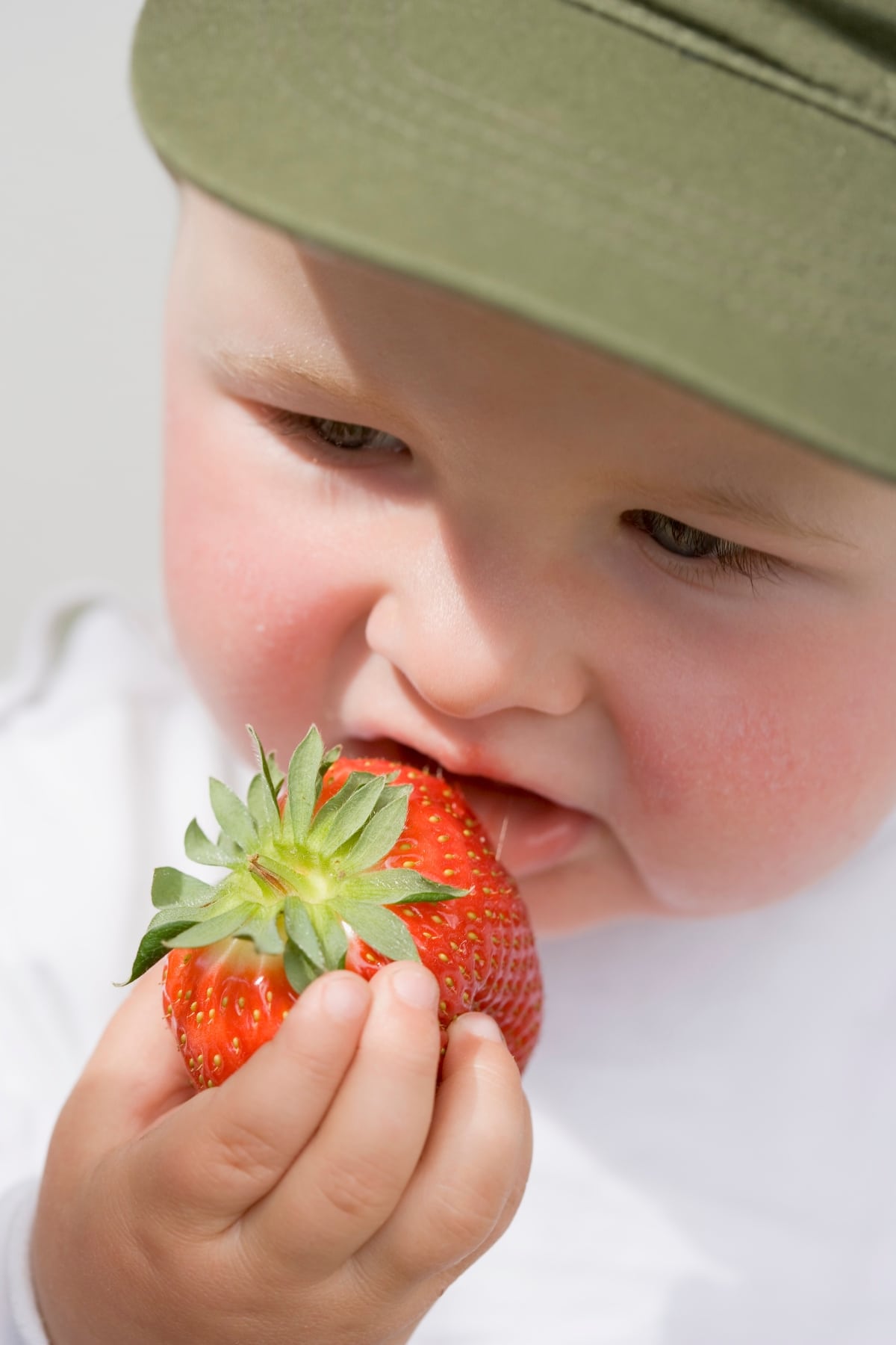 baby wearing a green cap eating a whole strawberry.