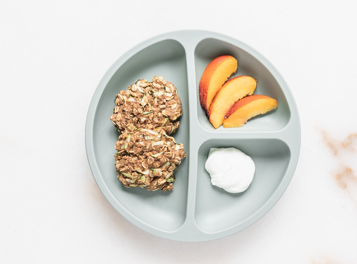 green baby plate with two zucchini banana oat breakfast cookies, peach slices, and yogurt.