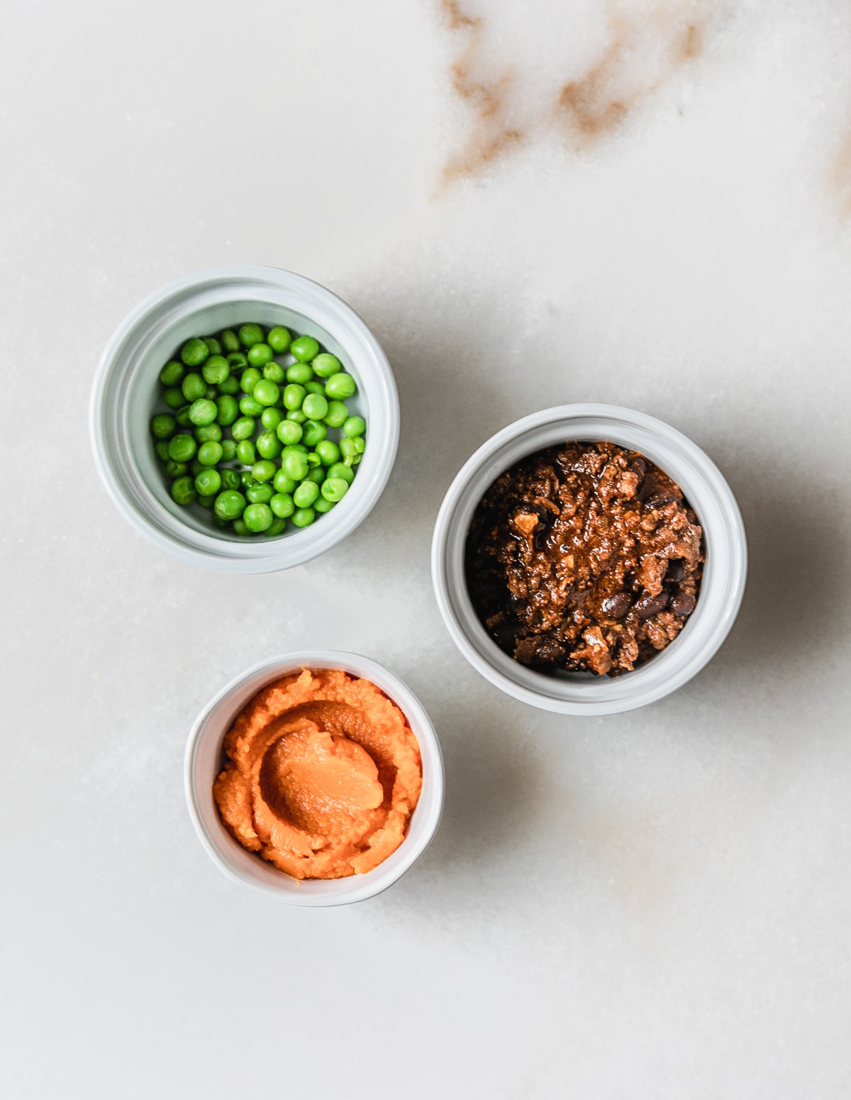 three small bowls of peas, pumpkin puree and chili on a white background.