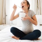 pregnant woman on a bed in a grey shirt eating from a container with a spoon