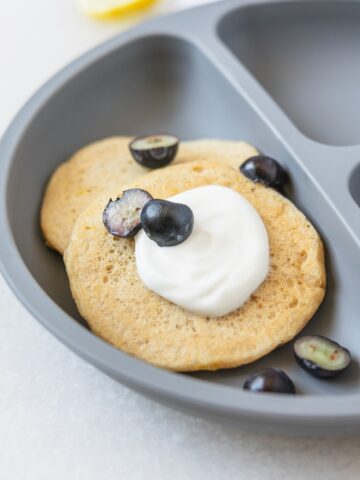 two baby ricotta pancakes topped with yogurt and blueberries on a grey baby plate.