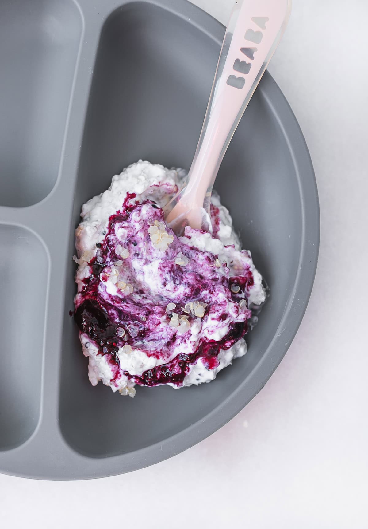 quinoa mixed with yogurt and blueberry compote on a grey baby plate with a pink baby spoon.