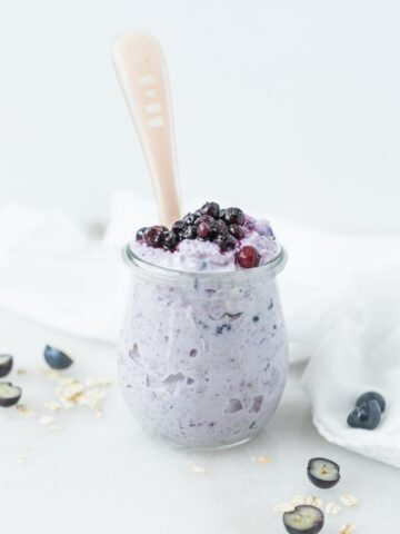glass jar with blueberry overnight oats for baby topped with thawed frozen blueberries and a spoon in it.