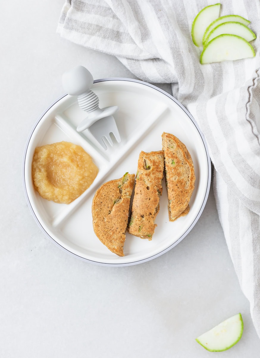 baby zucchini bread pancake cut into strips and applesauce on a baby plate with a baby fork.