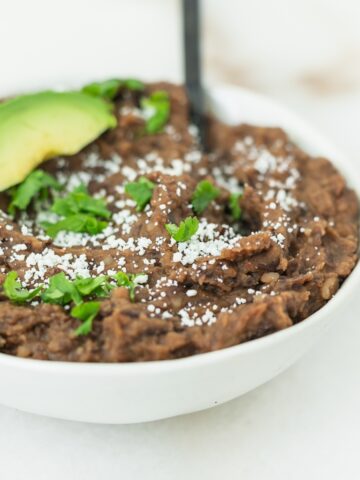 easy refried black beans in a white bowl topped with cotija cheese, cilantro and an avocado slice.
