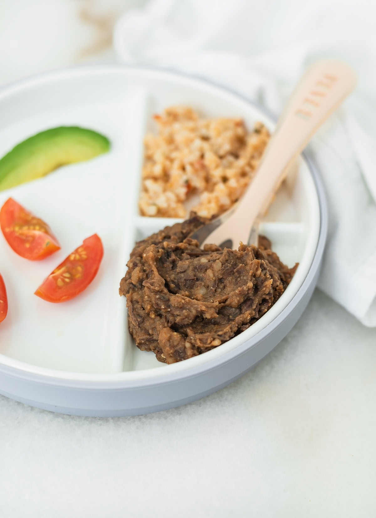 refried black beans on a divided baby plate with rice, tomato quarters and avocado slice.