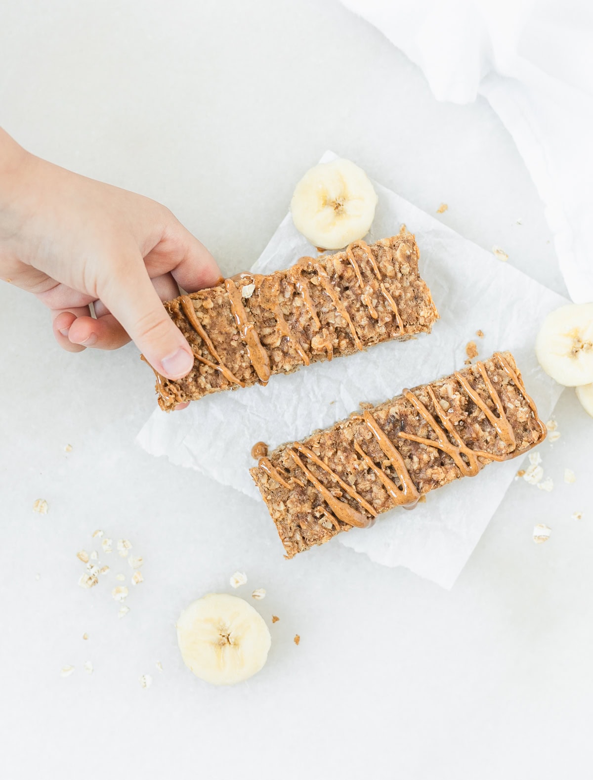 two soft baked oatmeal bars surrounded by banana slices with a toddler hand reaching to pick up a bar.