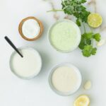 3 different tahini sauces in small bowls surrounded by herbs, lemon and lime halves, garlic and yogurt.