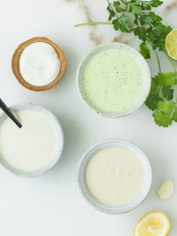 3 different tahini sauces in small bowls surrounded by herbs, lemon and lime halves, garlic and yogurt.