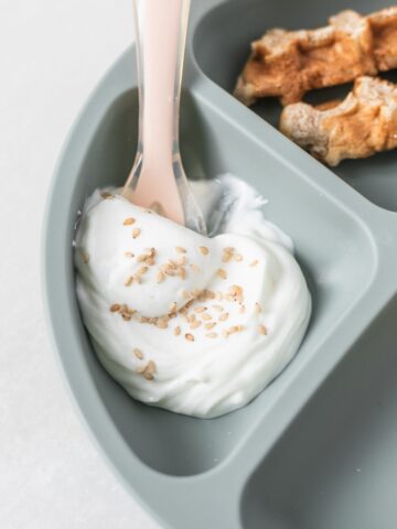 sesame seeds sprinkled on yogurt in a section of a baby plate with a pink baby spoon.
