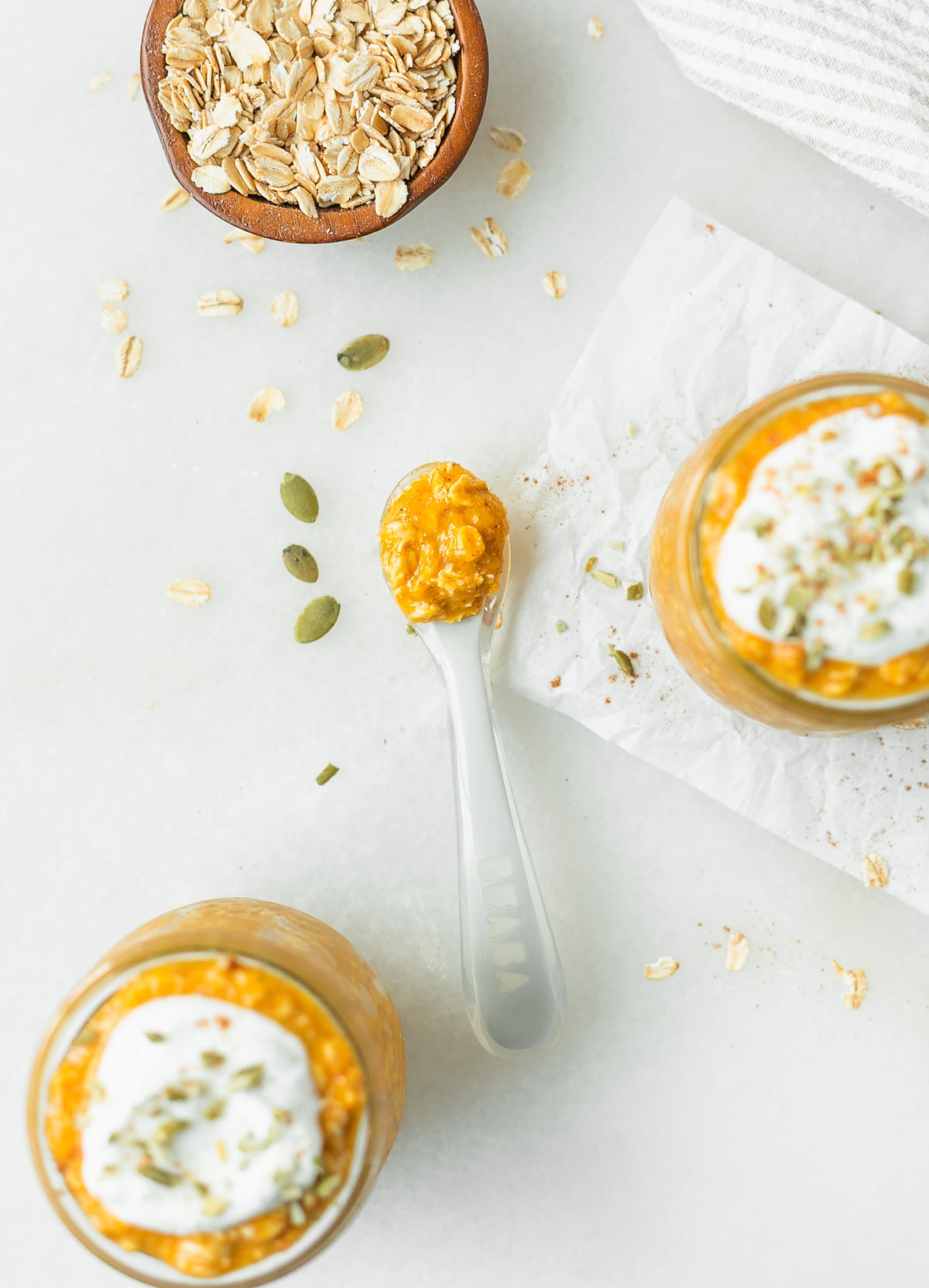 overhead view of pumpkin spice overnight oats on a baby spoon surrounded by pumpkin seeds, containers of overnight oats, and dry oats.